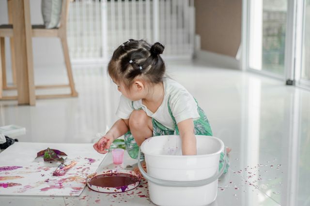 girl reaching into a white bucket, with paint all over her hands and the floor around her