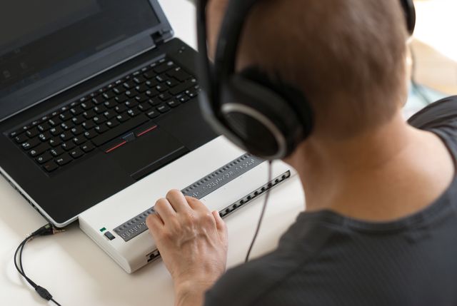 Over the shoulder, a person wearing headphones, using a braille display attached to a laptop